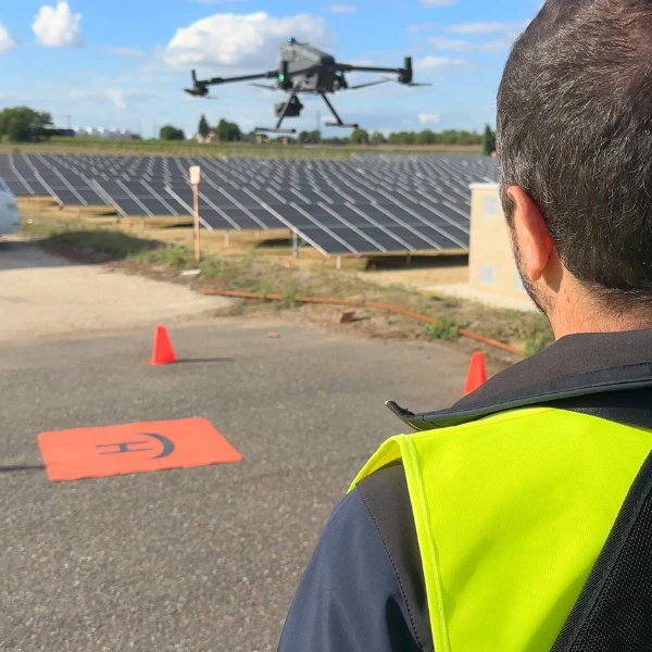 Alumno con chaleco reflectante observando desde el suelo el vuelo de un dron industrial sobre hileras de paneles solares y plataforma de aterrizaje portátil durante el curso de inspección de plantas fotovoltaicas de Toledrón.