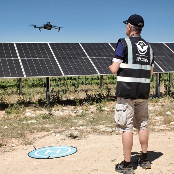 Piloto con chaleco corporativo de la escuela Toledrón controlando un dron industrial en vuelo sobre hileras de paneles solares junto a helipuerto portátil de despegue durante prácticas de inspección fotovoltaica.