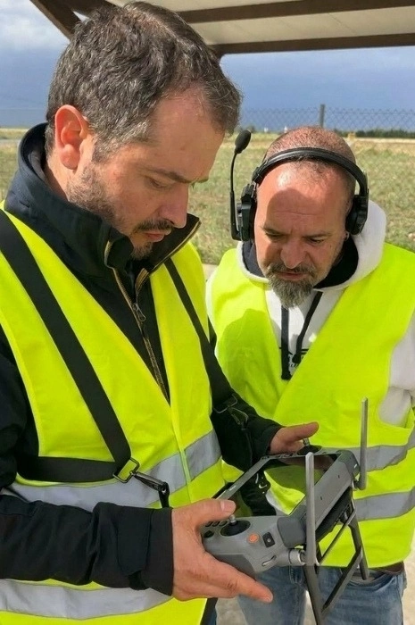 Instructor de Toledrón con cascos de comunicación guiando de cerca a un alumno con chaleco reflectante que maneja la emisora inteligente con visera de protección para el curso de drones.