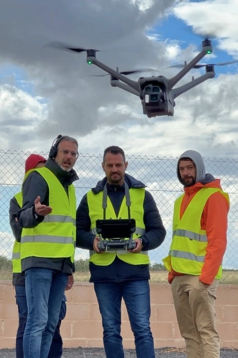 Tres alumnos con chalecos reflectantes y piloto con emisora de control inteligente realizando instrucción técnica de vuelo y coordinación por radio mientras un dron industrial vuela en el cielo para Toledrón.