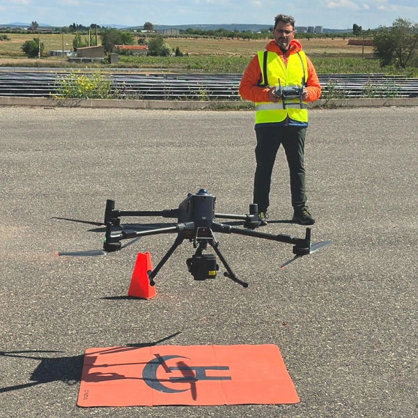 Instructor de vuelo con chaleco de alta visibilidad operando la emisora con pantalla de un dron profesional industrial sobre helipuerto portátil de despegue durante prácticas de campo en Toledo.