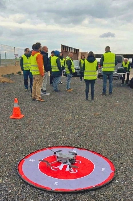 Dron multirrotor sobre helipuerto portátil de despegue con rosa de los vientos y cono de balizamiento en primer plano frente a grupo de alumnos con chalecos reflectantes en las prácticas del curso de Toledrón.