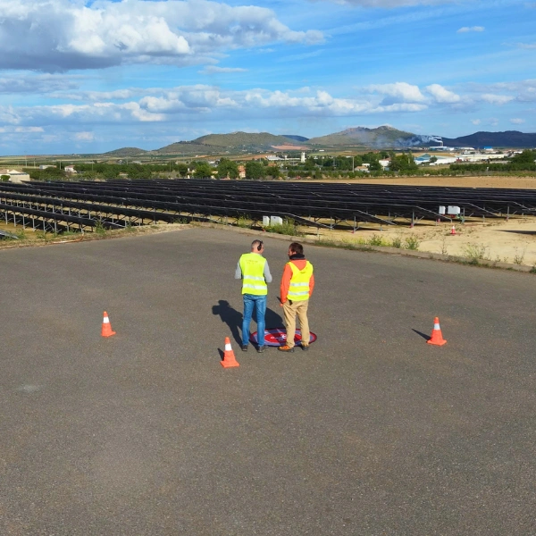 Instructor y alumno con chalecos reflectantes y protectores auditivos realizando prácticas de vuelo con dron en helisuperficie portátil junto a una planta solar para el curso de inspección fotovoltaica de Toledrón.