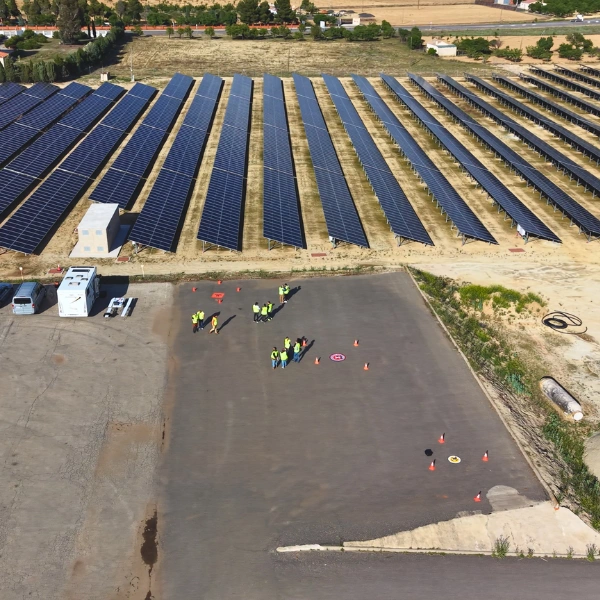 Perspectiva aérea cenital de un parque solar fotovoltaico con hileras de paneles y explanada de prácticas donde instructores y alumnos con chalecos reflectantes realizan maniobras de vuelo con dron en Toledo.