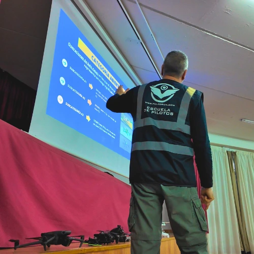 instructor-explicacion-teoria-drones-aula Instructor de la escuela de pilotos de drones Toledrón señalando una presentación teórica proyectada en pantalla durante un curso en aula.