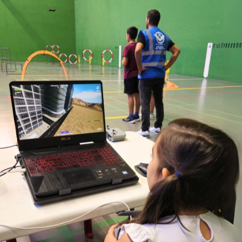 ninos-volando-drones-simulador-fpv Niña utilizando un simulador de drones en portátil mientras instructores enseñan a otros niños en un circuito FPV con aros iluminados en un polideportivo.